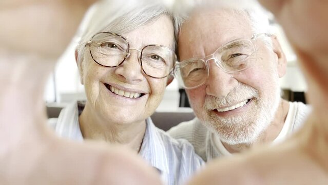 Close up portrait happy sincere middle aged elderly retired family couple making heart gesture with fingers, showing love or demonstrating sincere feelings together indoors, looking at camera.
