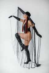 A red-haired girl in a black bodysuit, patent leather boots, gloves and a hat with threads stands...