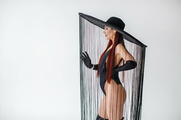 A portrait of a red-haired girl in a black bodysuit, gloves and a hat with threads stands in the...