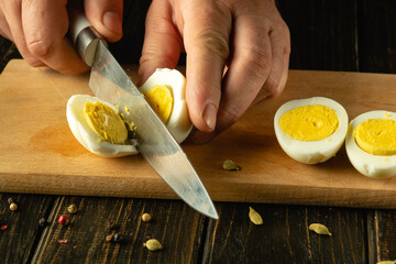 Close-up of a chef hands with a knife slicing a boiled egg on a kitchen cutting board before...