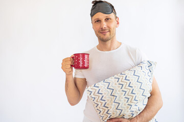 portrait of a young man in pajamas holding a pillow and a cup isolated on white background