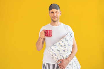 portrait of a young man in pajamas holding a pillow and a cup isolated on yellow background