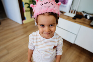 Portrait of a young smiling girl with a happy birthday cap on her head. A happy child is standing in the children's playroom and looking at the camera. High angle view.