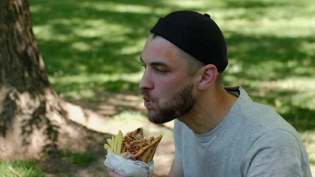 Traditional Greek street food - chicken gyros with sauce, vegetables and French fries wrapped in pita. Young guy eats a gyros in spring park on sunny day. Middle eastern street food cuisine