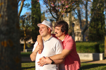 Fototapeta premium Couple of gay men hugging standing and smiling looking in front with sunset light giving them in a park with nature in the background. LGBT love concept