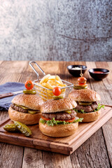 Three burger sliders placed on a wooden board in a rustic atmosphere. Very tasty burgers with melted cheese, lettuce, tomato, potatoes and sauce