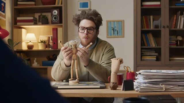 Medium over-shoulder shot of young male office buffoon harassing colleague in suit arriving for work in morning, making faces, playing with wooden dolls and hand, trying to attract attention