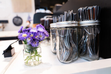 Small bright purple flowers in a glass bottle along with a spoon and fork are placed on a table...