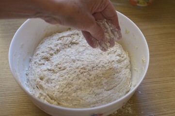 hand adds flour onto raw yeast dough in a ceramic bowl on a wooden kitchen table.