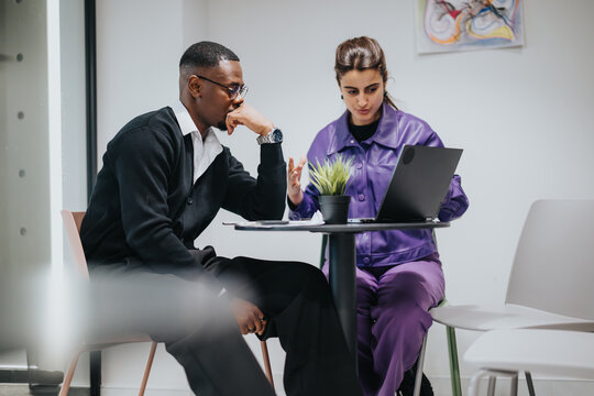 An African American man and an Indian woman, dressed in business attire, collaborate on a project using a laptop in a brightly lit office space.