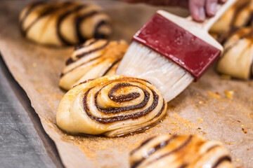Person glazing raw cinnamon rolls with a pastry brush and preparing them for baking