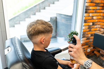 A boy sits in a barbershop while he is sprayed with water to start a haircut. A master in a...