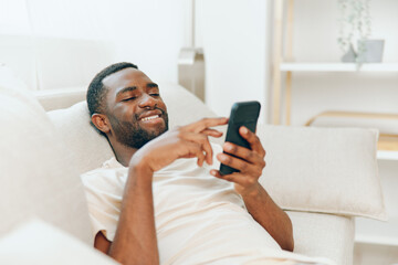 Happy African American man sitting on a black sofa, typing a message on his mobile phone He is relaxing at home, enjoying the modern lifestyle and wireless technology The cozy apartment provides a