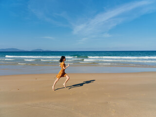 Woman in a vibrant yellow dress joyfully running along the sandy beach shore with the vast ocean in...