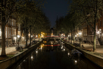 Houses and bridges reflecting on the calm waters of the canals in Amsterdam at night with cars...