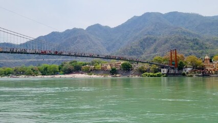 Locked shot showing the majestic suspension bridge Ram Lakshman jhula going over blue waters of Ganga river with people crossing and the Himalaya mountains in the background