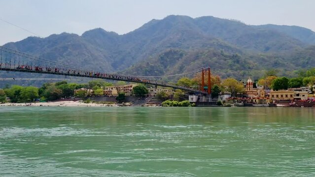 Panning shot showing the majestic suspension bridge Ram Lakshman jhula going over blue waters of Ganga river with people crossing and the Himalaya mountains in the background