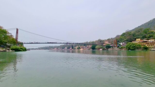 mid river shot showing the majestic suspension bridge Ram Lakshman jhula going over blue waters of Ganga river with people crossing and the Himalaya mountains in the background