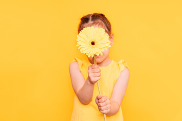 Little girl covering her face with a flower in front of a yellow background.