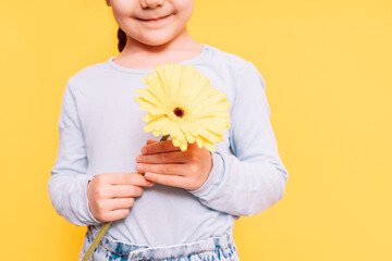 Child holding flower in her hands on Mother's Day holiday against yellow background.
