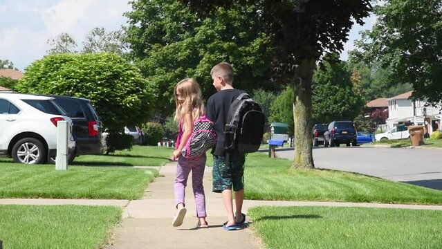 Two young children are wearing book bags and walking to school on a suburban sidewalk outside on a sunny day with backpacks for an back-to-school or safety concept.