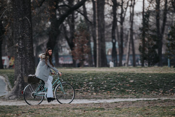 A business lady in a chic coat rides her bicycle through a tranquil urban park setting.