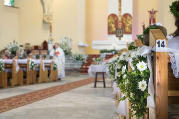 Interior of Catholic church decorated with white flowers and ribbion during First Holy Communion celebration, in background blurred priest and altar