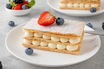 Traditional French dessert millefeuille with vanilla cream and fresh berries on a white plate on a gray concrete background.