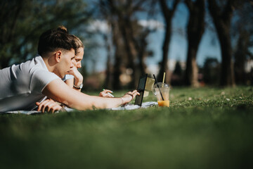 A young school couple lying on the grass together, focused on a tablet screen against a backdrop of...