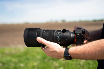 A close-up photo capturing a person's hands adjusting a large black camera lens outdoors, with a blurred green field background.