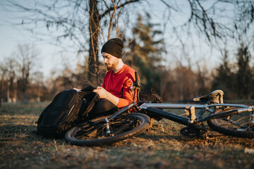 A young man takes a peaceful break, lying on the ground beside his bicycle, writing in his notebook while sitting in a serene park setting.