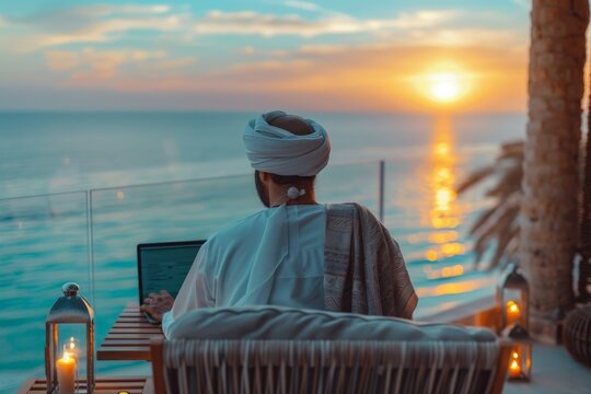 Tranquil scene of a man in cultural Arabian clothing working on his laptop during a seaside sunset
