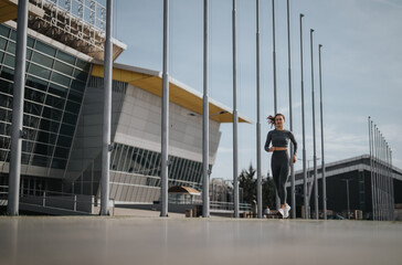 Fitness-focused woman in sportswear running near contemporary buildings on a clear day, showcasing...