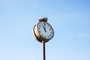 A quartz clock on a metal pole against a blue sky reads 1155