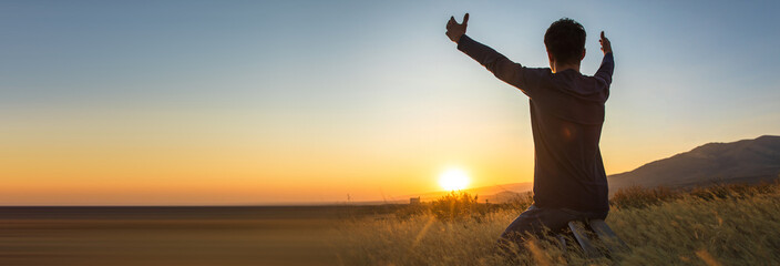 Boy kneeling praying with arms outstretched at sunset, spiritual concept, stock photo