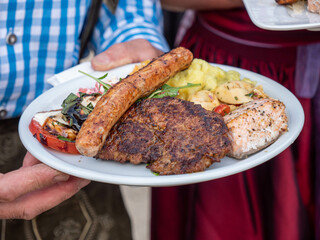 A man in traditional Bavarian costume holds a barbecue plate with bratwurst, grilled steak and various salads