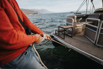 Close-up of a man securing a pontoon boat with a thick rope, preparing for a ride on a tranquil lake surrounded by mountains. - obrazy, fototapety, plakaty