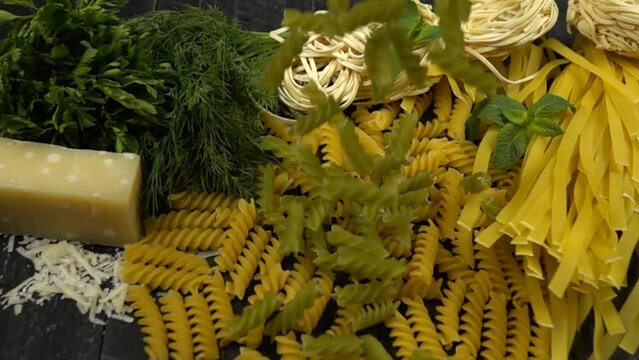 Green pasta is poured on the table, slow motion.  Different kind of pasta, cheese parmesan, bunches fresh dill, parsley are on a black table. 