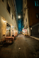 Fototapeta premium In a quaint narrow street at night, tables and chairs are set up under the dim glow of streetlights. Customers enjoy dining, chatting, and sipping drinks al fresco