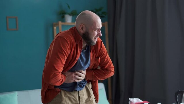 Bearded man suffering with severe stomach pain at home. Male hands holding abdomen suffering from ache and indigestion problem. painful sensation in belly