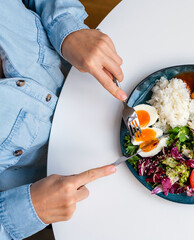 Directly above plate with a healthy and balanced meal and female's hands holding folk and knife. Rice, egg, vegetables, fish.