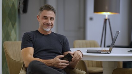 Confident mature man smiling at camera, sitting with laptop in cozy room. Businessman working at home with computer. Happy mid adult man with smile. Entrepreneur managing business online.