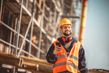 Construction site manager standing  wearing safety vest and helmet,using his phone at construction...