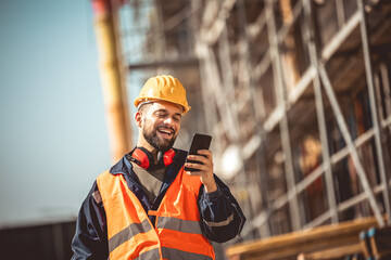Construction site manager standing  wearing safety vest and helmet,using his phone at construction site.