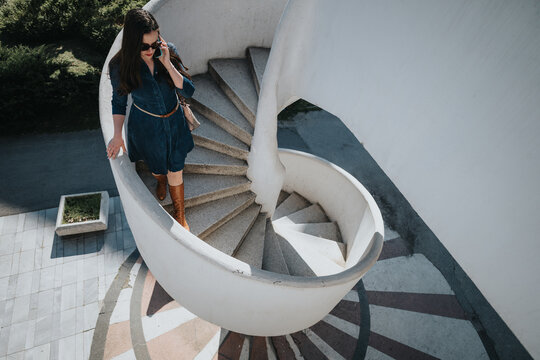Confident businesswoman in a denim dress talking on the phone on a sunny day, as she walks down an exterior spiral staircase