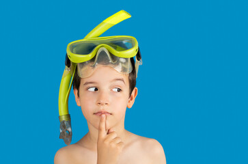 Pensive kid with snorkel mask tuba and snorkel looks away at copy space thinking isolated on a blue background, kid lips hold finger near mouth.Snorkeling, swimming, vacation concept.