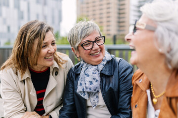 Three mature retired women laughing while talking sitting outside. Small group of mature females having fun enjoying a conversation bonding at city street. Elderly friendship concept.