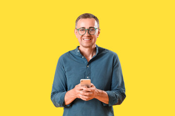 Mature man in eyeglasses using mobile phone on yellow background