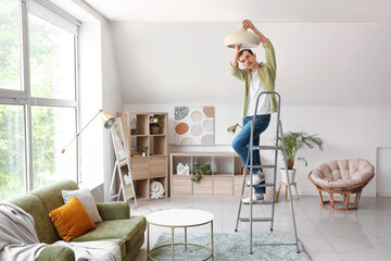 Young man standing on ladder and changing light bulb in lamp at home