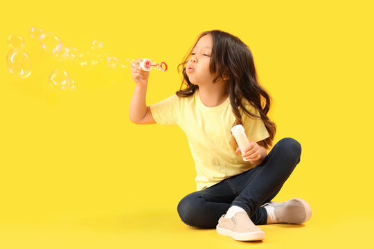 Cute little girl blowing soap bubbles on yellow background